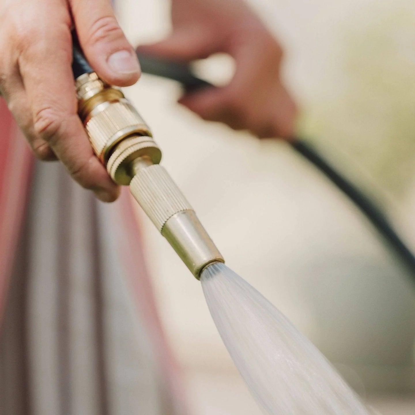 Person holding a brass garden hose nozzle with water spraying out, blurred background