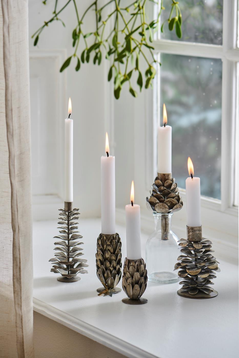 Five metal pinecone candle holders of varying heights and finishes holding lit white candles on a white windowsill, with mistletoe hanging above and a glass bottle among them