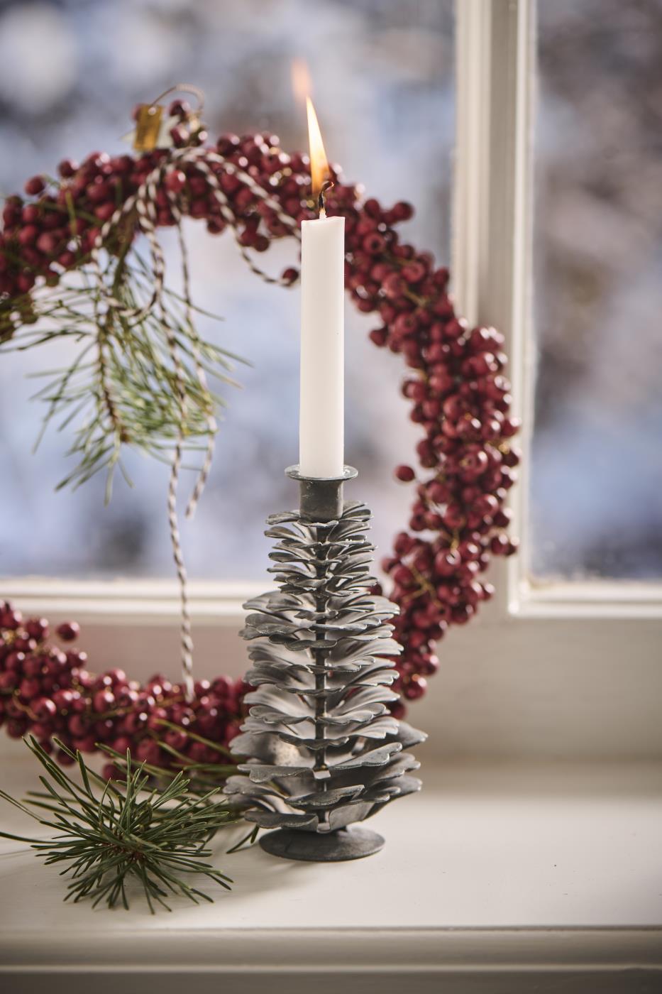 Close-up of a small grey metal pinecone candle holder with a lit white candle on a windowsill next to a red berry wreath