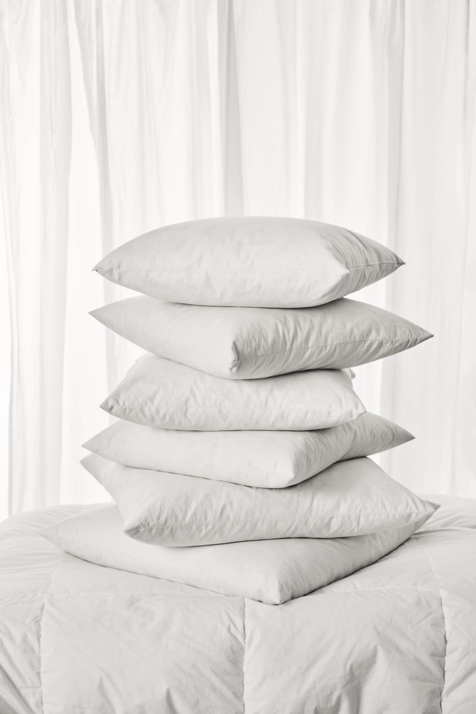 A neat stack of six white cushion fillers on a folded white quilt against a sheer white curtain