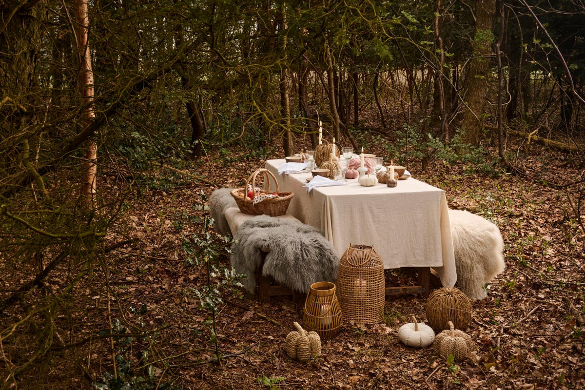 Woodland autumn table set with fur throws, a wicker basket, rattan lanterns, and seagrass and fabric pumpkins on the ground