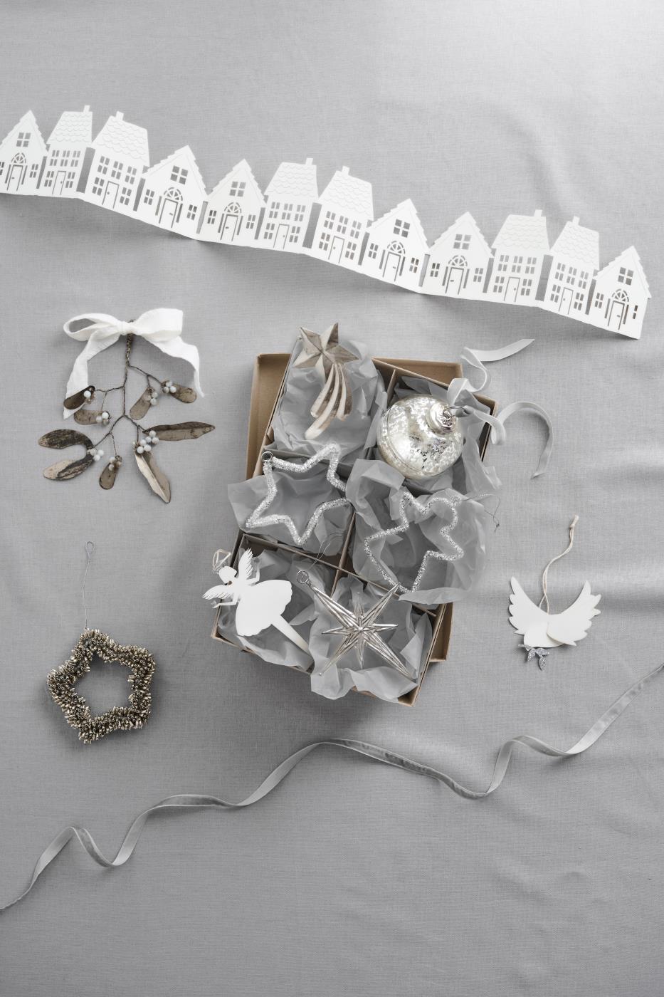 Assorted silver and white Christmas ornaments including stars, angels, a mercury glass bauble, and ribbon, arranged in a box beside a paper house garland
