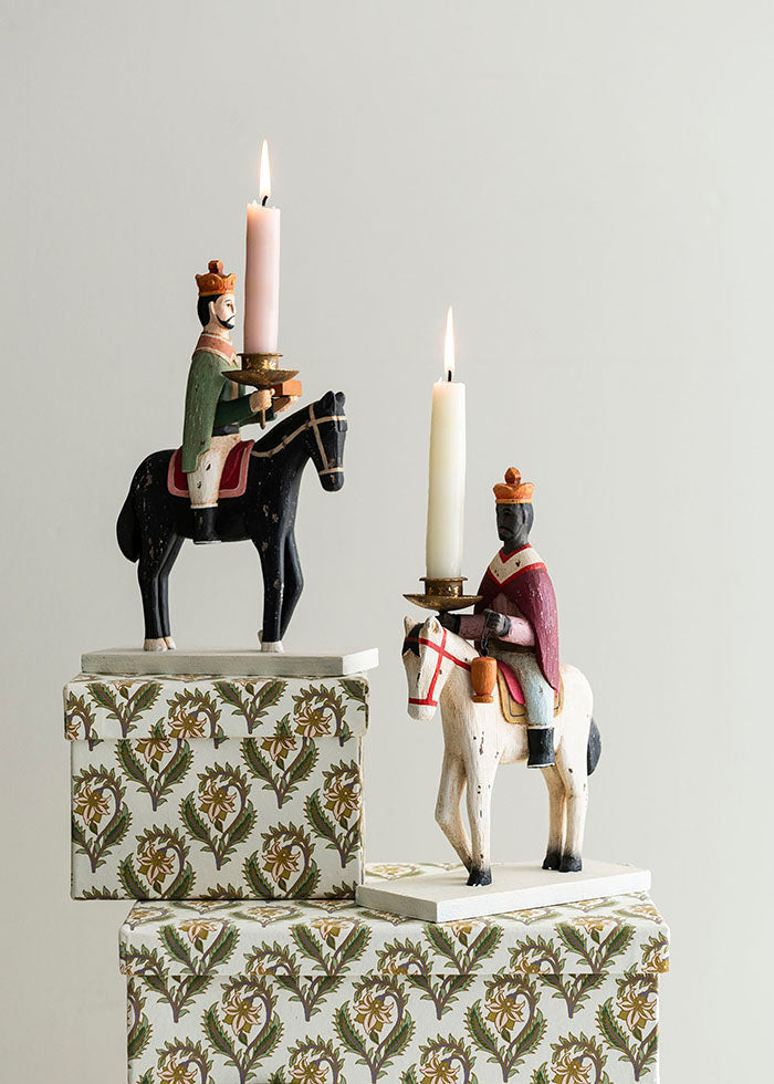 Decorative candle holders with figurines of men on horseback on patterned boxes against a gray background.