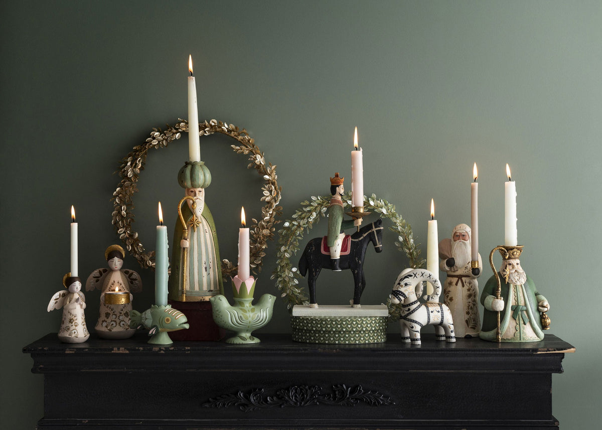 A decorative holiday mantel display featuring multiple ceramic candle holders, lit taper candles, a green wreath, and figurines including Santa Claus, an angel, a horse rider, and a white dog, set against a dark green wall.