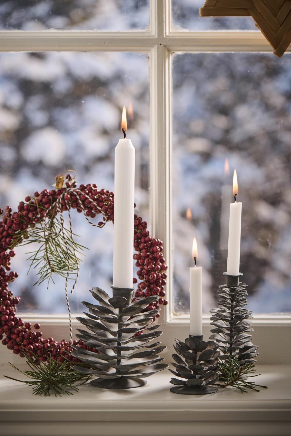 Grey metal pinecone-shaped candle holders in three sizes with lit candles on a windowsill beside a red berry wreath, snowy view outside