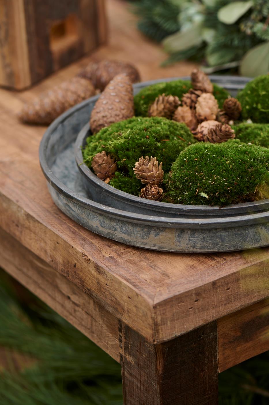 Round weathered metal tray styled with moss and pinecones as a Christmas table centrepiece on a rustic wooden table