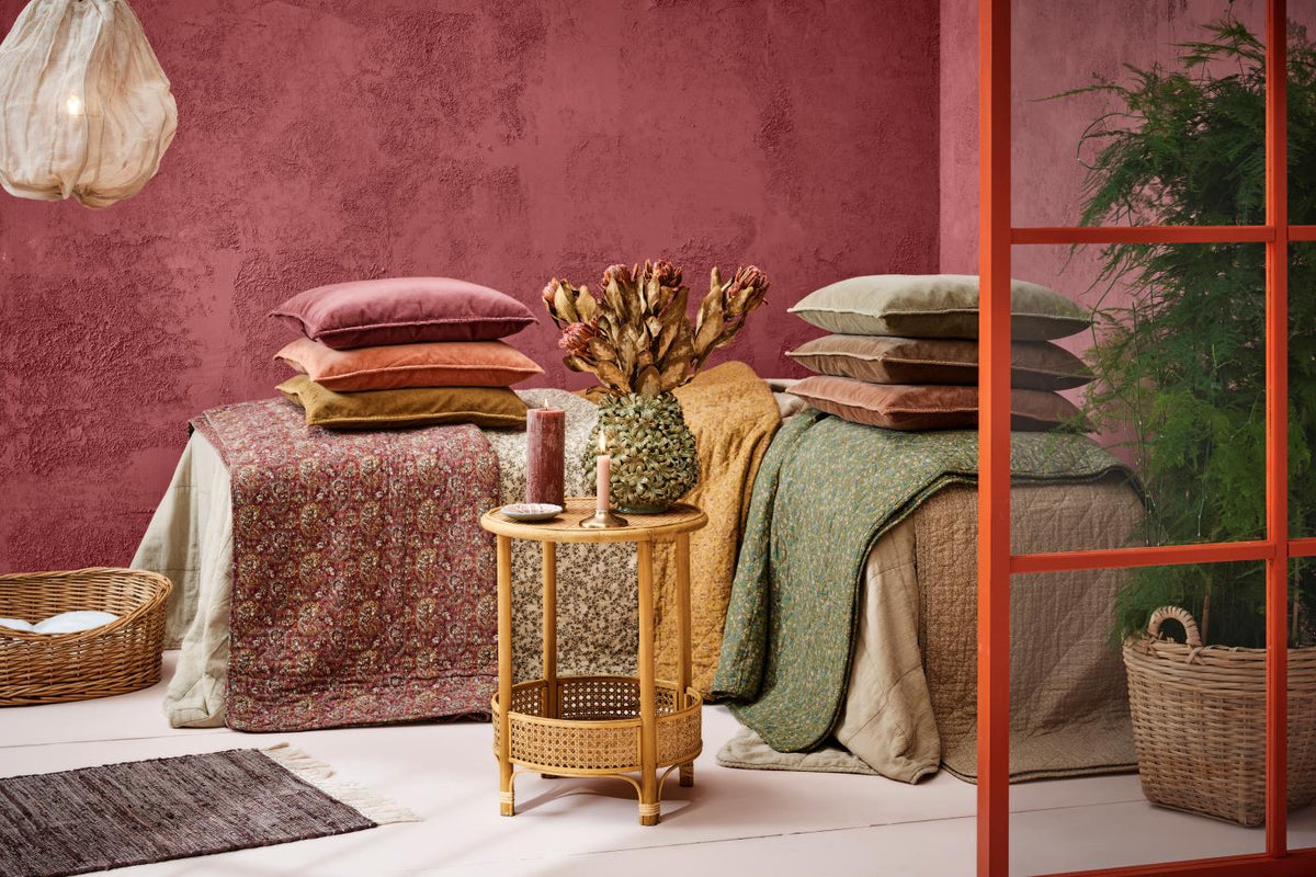 Stacks of velvet cushions in pink, terracotta, ochre, and green tones on quilted throws and a floral bedspread against a rose-coloured textured wall